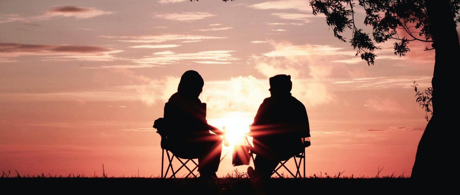 Sunset photo of two people seated in lawn chairs on grass facing towards sunset while in conversation.