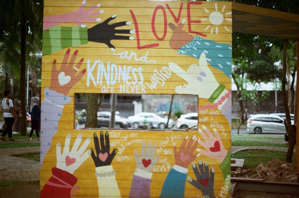 Large wood art piece painted yellow with multicolor hands with hearts in the palm, and the following word: Love and Kindness are Never Wasted"