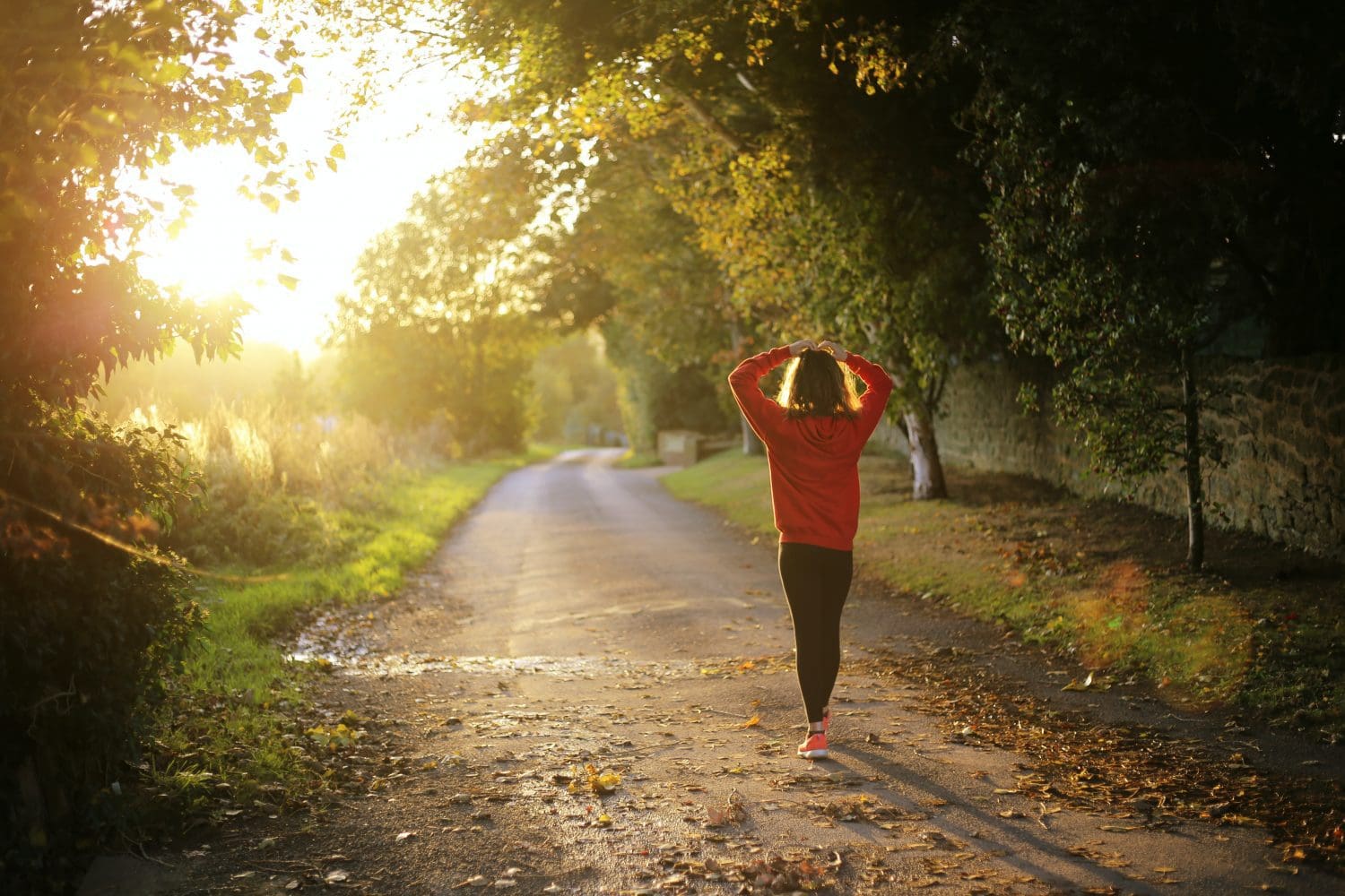 Woman walking along unpaved path towards sunrise with back to viewer